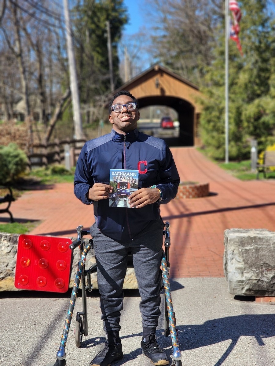 Young African American man with a walker smiling proudly outside in a park holding a book about trains.