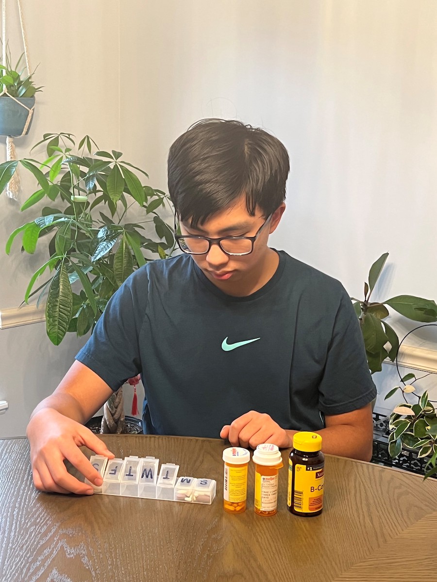 Young Asian male sitting at a table and organizing his medications with a pill organizer.