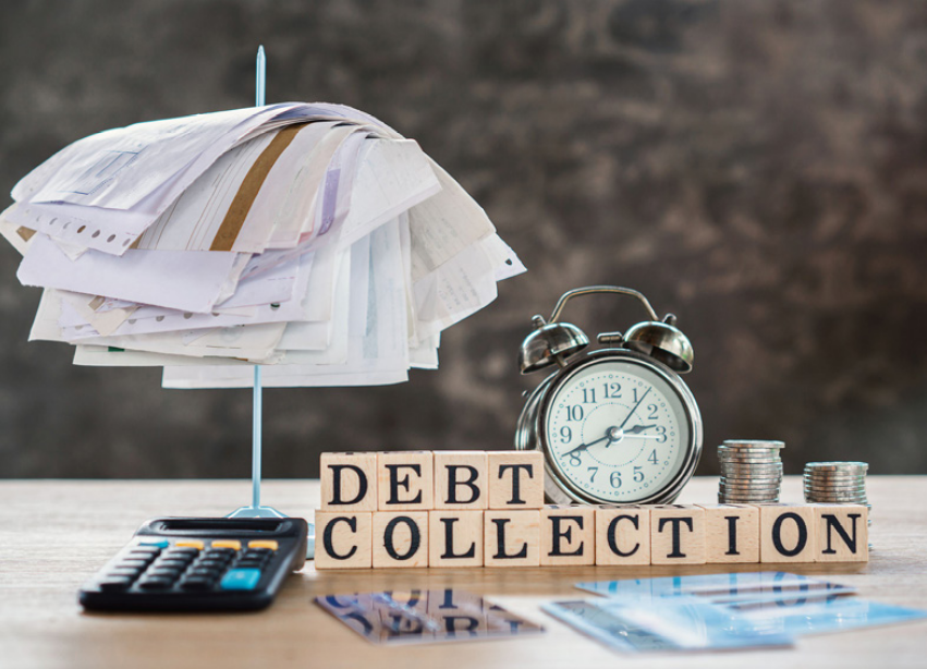 Multiple items on a table, including bills, coins, an alarm clock, a calculator, and wooden blocks with letters on them that ready out debt collection.