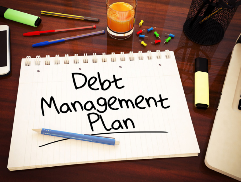 Multiple pens and a notebook on a desk. On the notebook there is writing that says debt management plan.