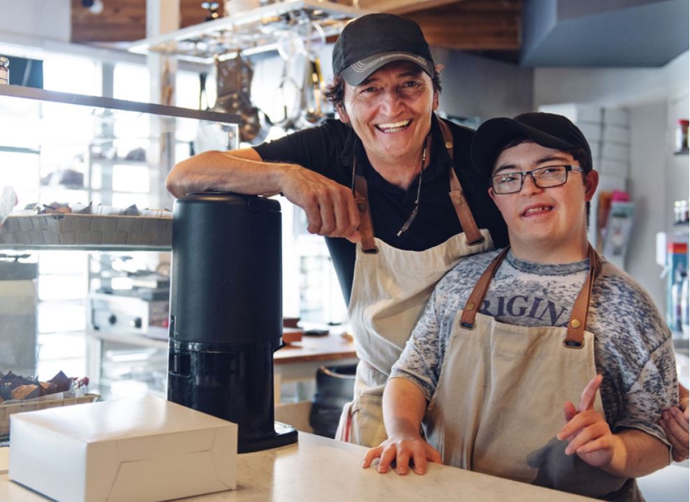 Two waiters in aprons smiling while behind the counter of a restaurant