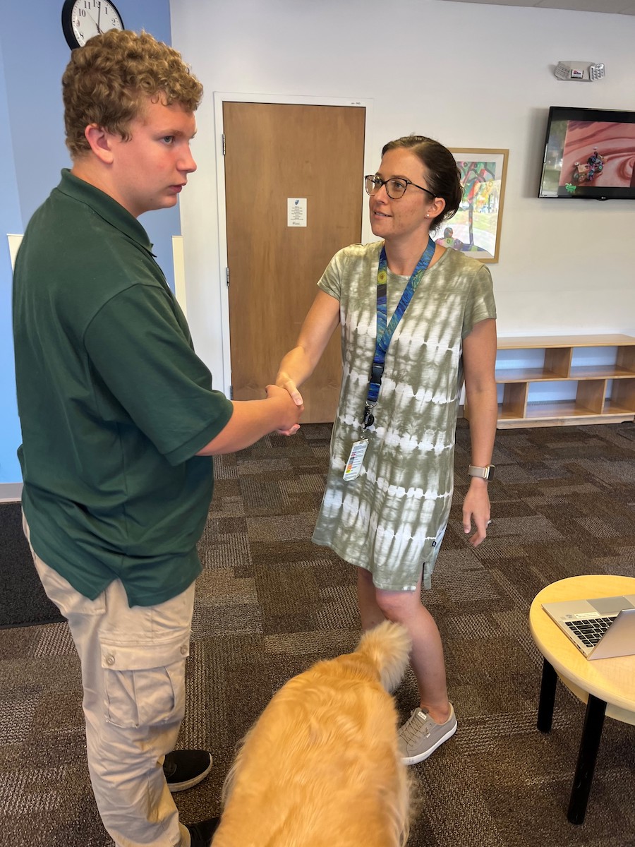 A young white male with his service dog shaking hands with a female doctor in a doctor’s office waiting area.