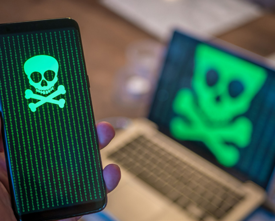 A laptop and a person holding a cell phone. Both screens have images of a skull and cross bones in front of a string of binary code.