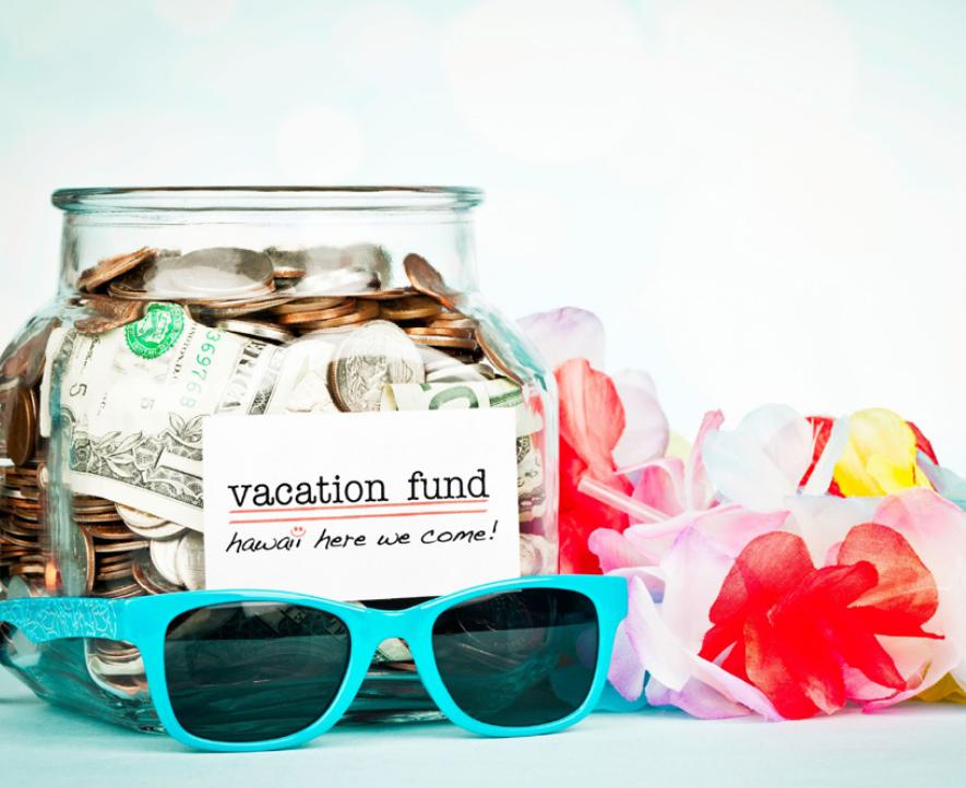 Glass jar with dollar bills in it. The jar has a label that says vacation fund. There are blue sunglasses and flowers next to the jar.