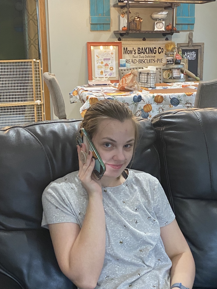 A young White woman sitting on a couch calling her doctor’s office on her cell phone.