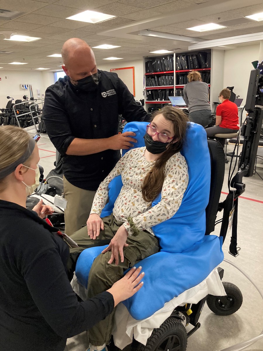 A White female in a wheelchair with two people assisting her with positioning.