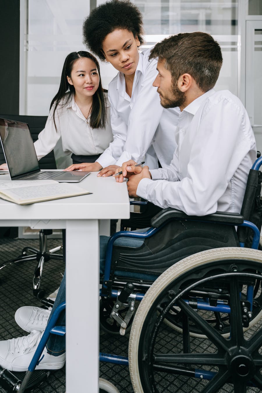 A White man in a wheelchair talking with an Asian and an African American woman looking at a computer screen.