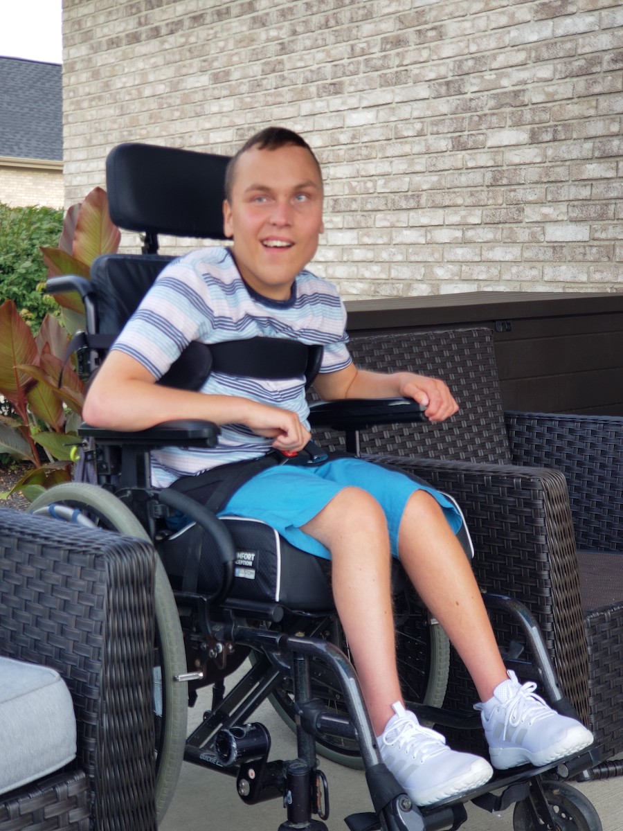 A young White man sitting outside in a wheelchair smiling.