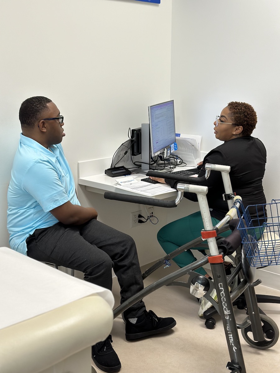 A young African American man with a walker sitting in a chair next to an African American healthcare provider taking notes on a computer.