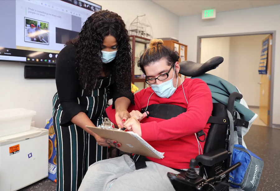 A White man in a wheelchair signing a document with an African American woman’s assistance.