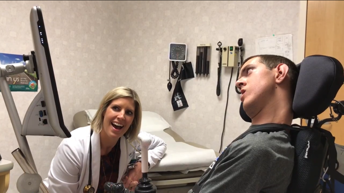 A young White man in a wheelchair using a communication device to talk with his White female doctor.