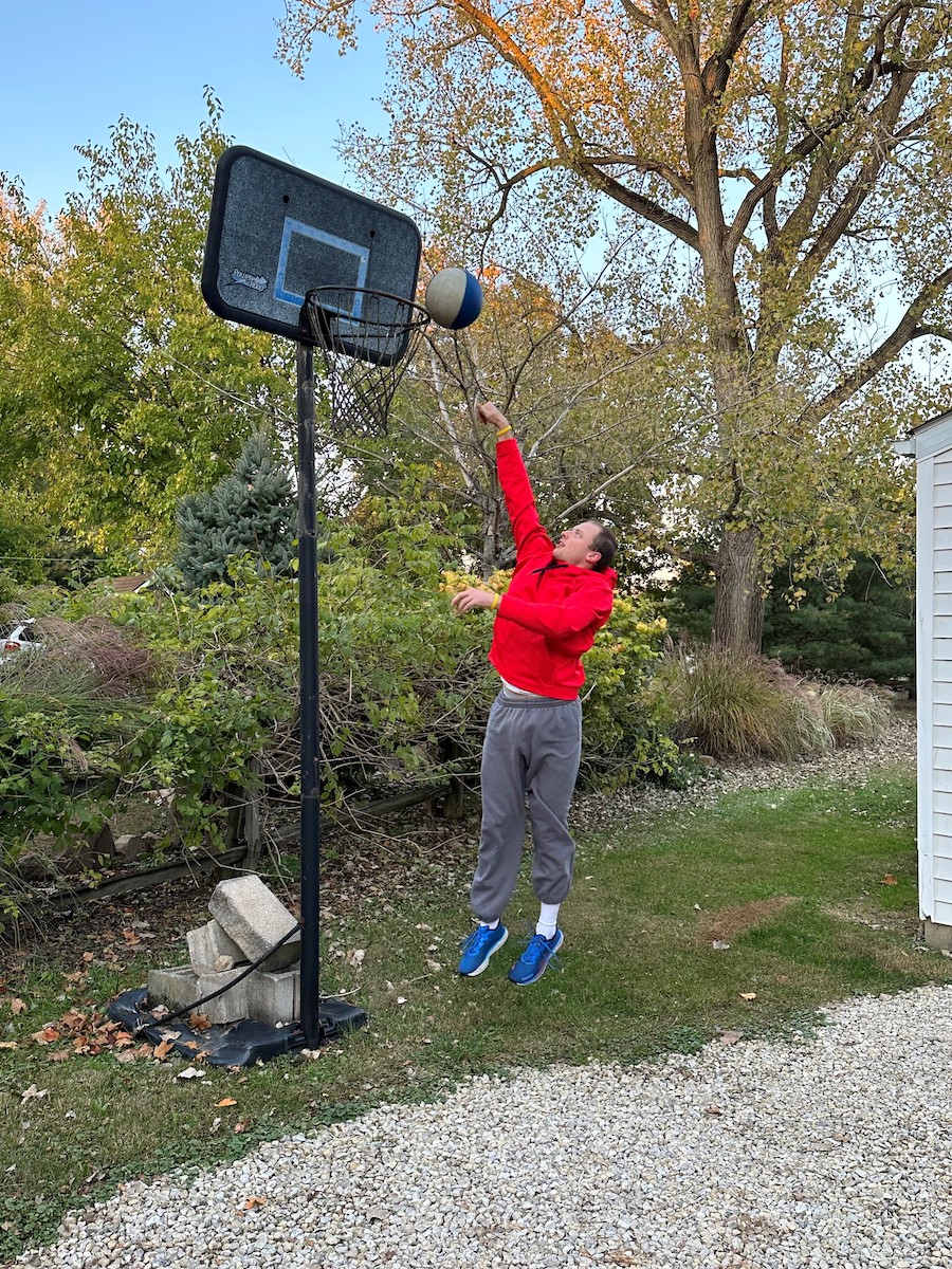 Young white male jumping up to shoot a basketball into the basketball hoop outside.