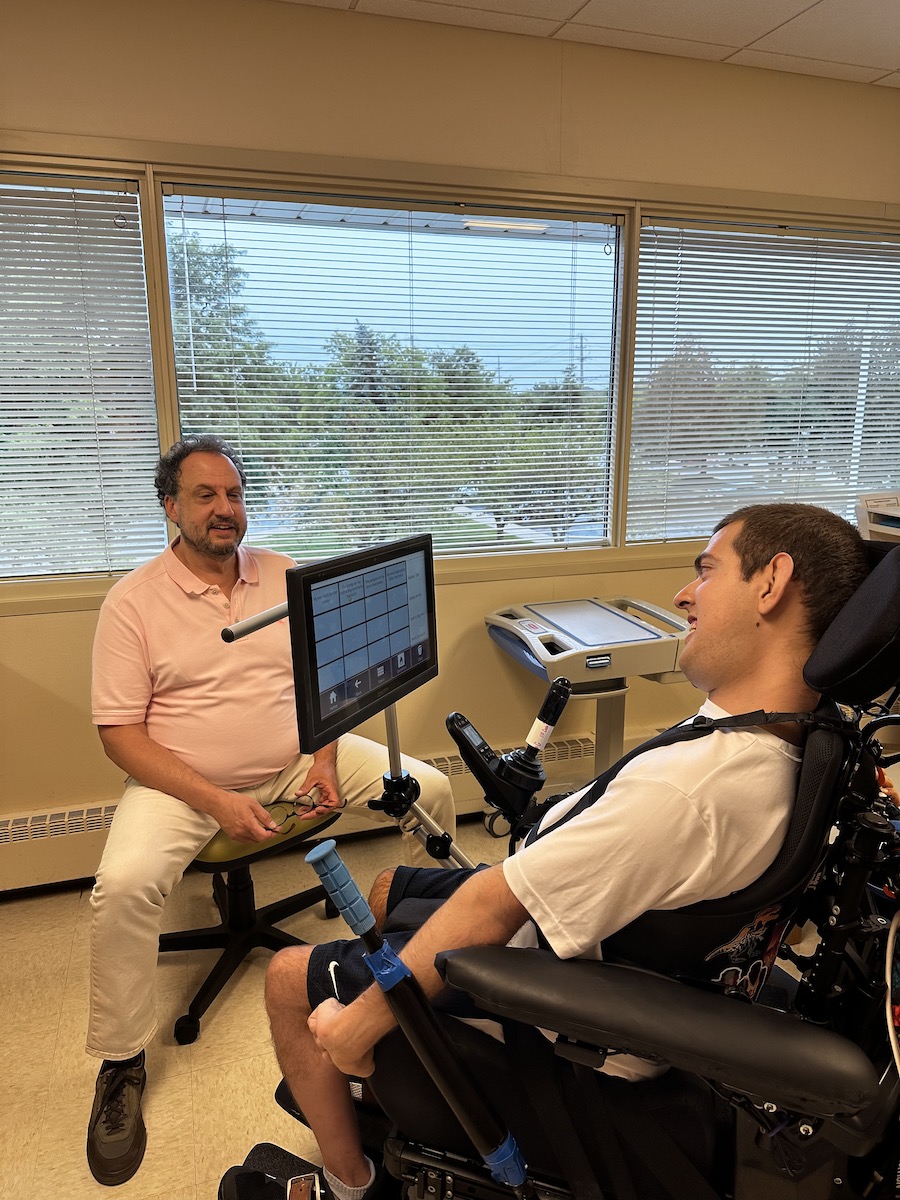 Young white man smiling in wheelchair using a communication device to talk with his doctor, who is sitting on a chair across from him.