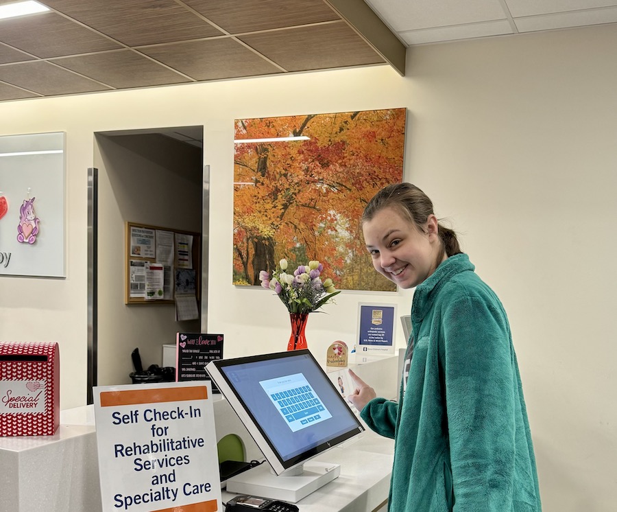 A young White woman checking in for a medical appointment at the doctor’s office, using a computer screen.