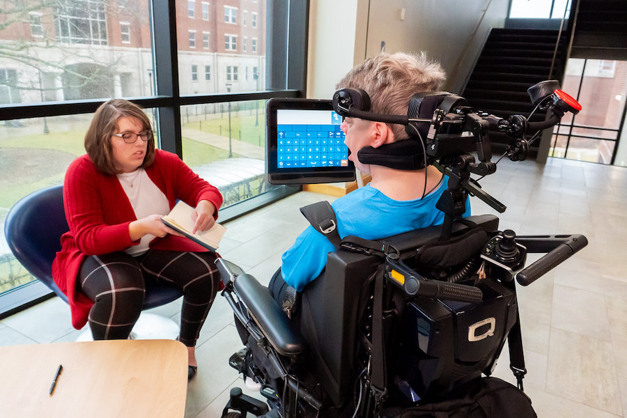 A White female in a wheelchair talking and reviewing notes with another White female sitting in a chair.